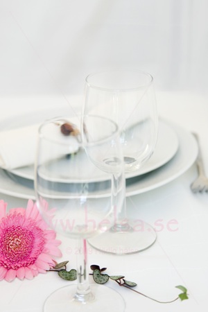 Vitt porslin, vita linneservetter, vinglas och bestick. Rosa gerbera samt hjärtan på tråd finns som dekoration på bordet. 